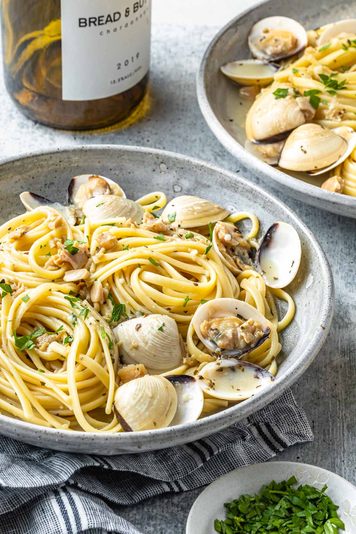 Clams and linguine in two bowls garnished with chopped parsley. A bottle of wine and a small bowl of chopped parsley next to the past bowls. 