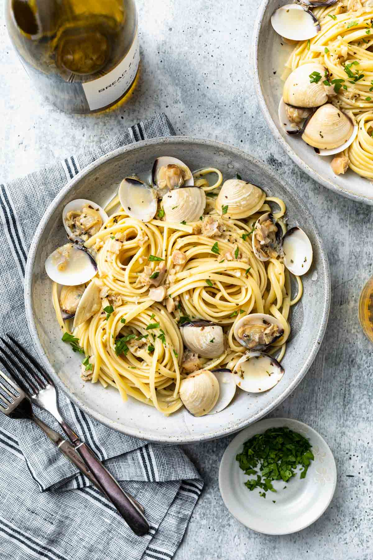 Overhead shot of a bowl of seafood pasta garnished with chopped parsley, and clam shells, with a bottle of wine and a bowl of parsley on the side. 
