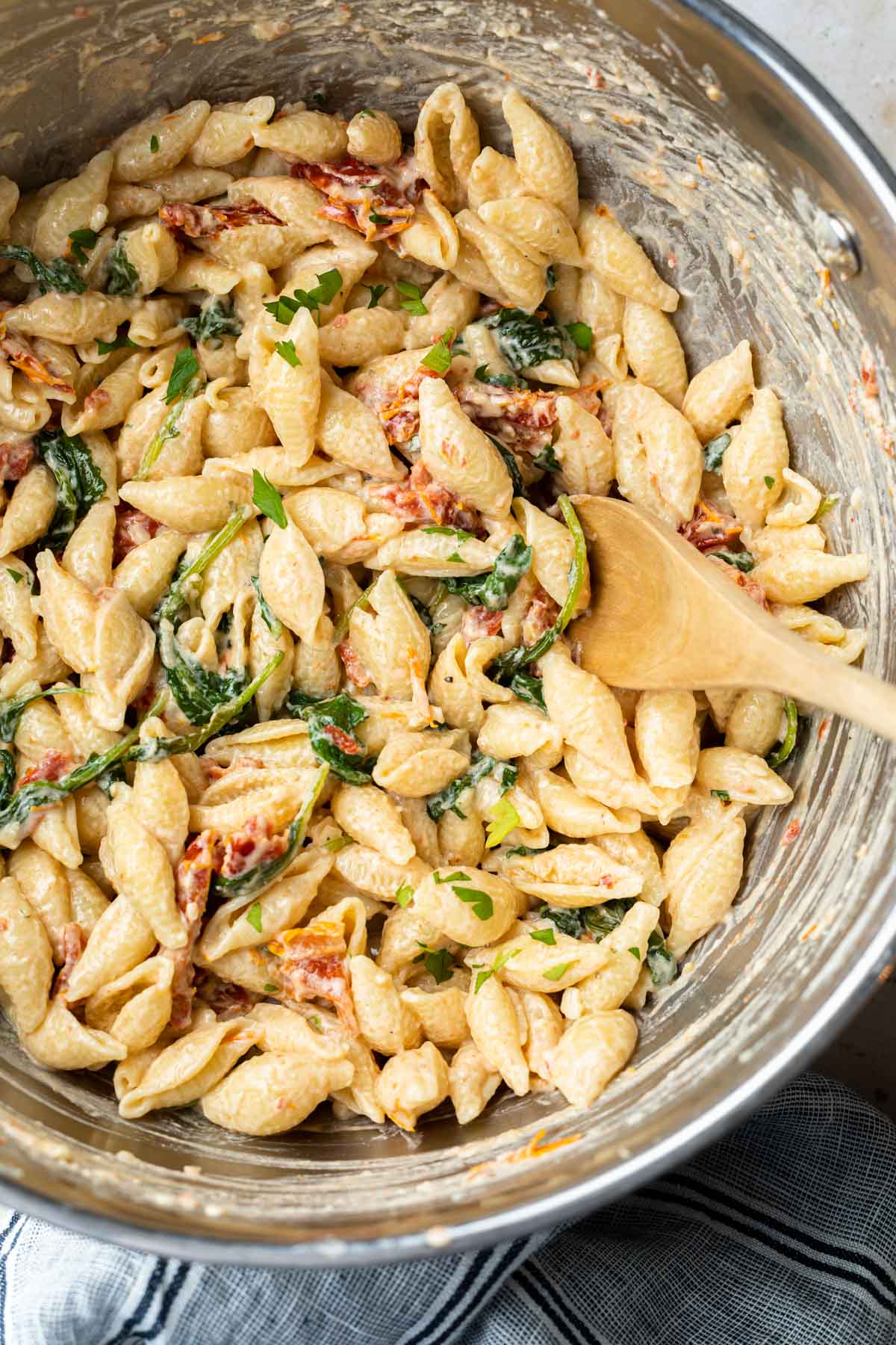 A close up of a stockpot of goat cheese pasta with sun-dried tomatoes and kale with a wooden spoon stirring. 
