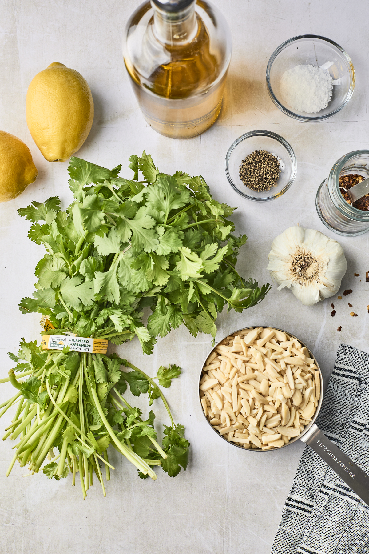 A bunch of cilantro, garlic, lemons, orzo, spices, grated cheese, a bottle of oil, and a striped cloth are arranged on a light surface—perfect for making fresh Cilantro Pesto.
