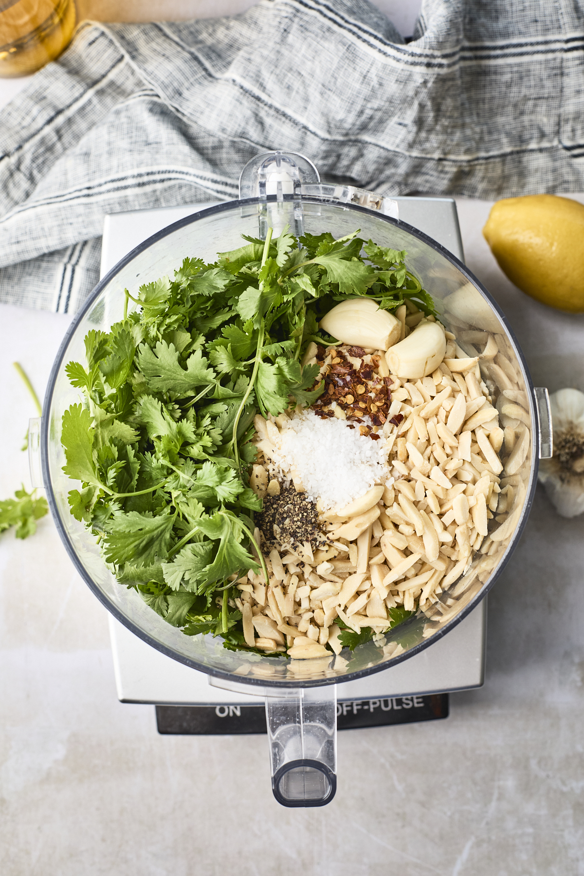 Top view of a food processor bowl filled with cilantro pesto ingredients—slivered almonds, garlic cloves, salt, pepper, and red pepper flakes—on a kitchen scale, with a lemon and kitchen towel nearby.