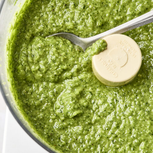 A close-up of a food processor filled with vibrant Cilantro Pesto sauce, with a spoon resting inside the fresh green mixture.