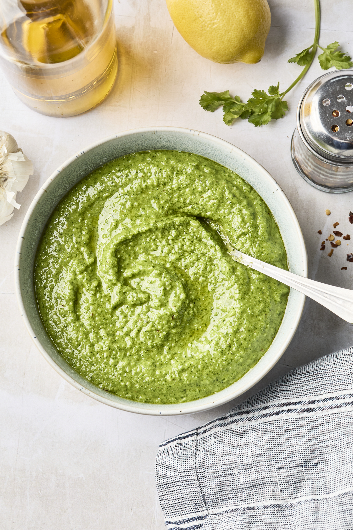 A bowl of Cilantro Pesto with a spoon inside, surrounded by a lemon, fresh cilantro, a shaker, glass container, garlic, and a striped cloth on a light surface.