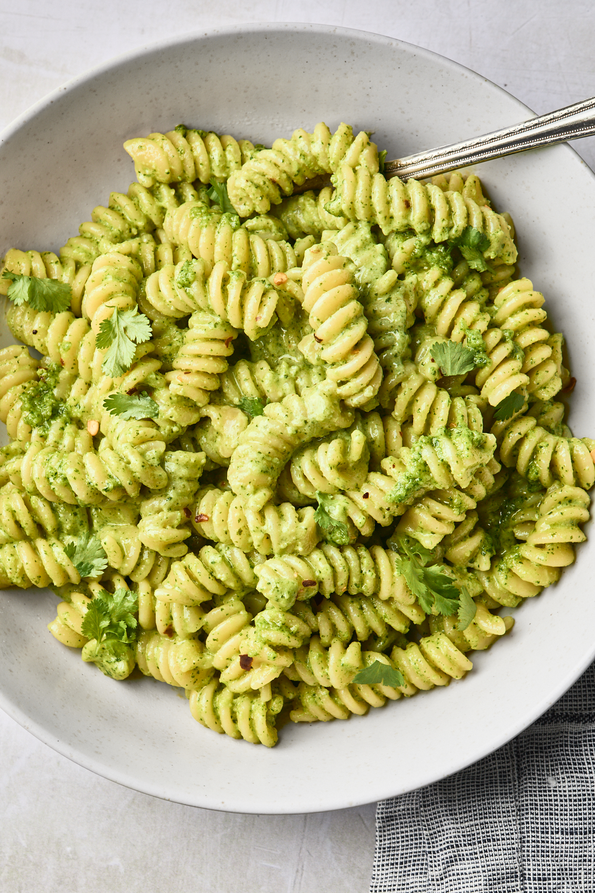 A white bowl filled with rotini pasta coated in vibrant cilantro pesto sauce, garnished with cilantro leaves, with a fork resting in the bowl.