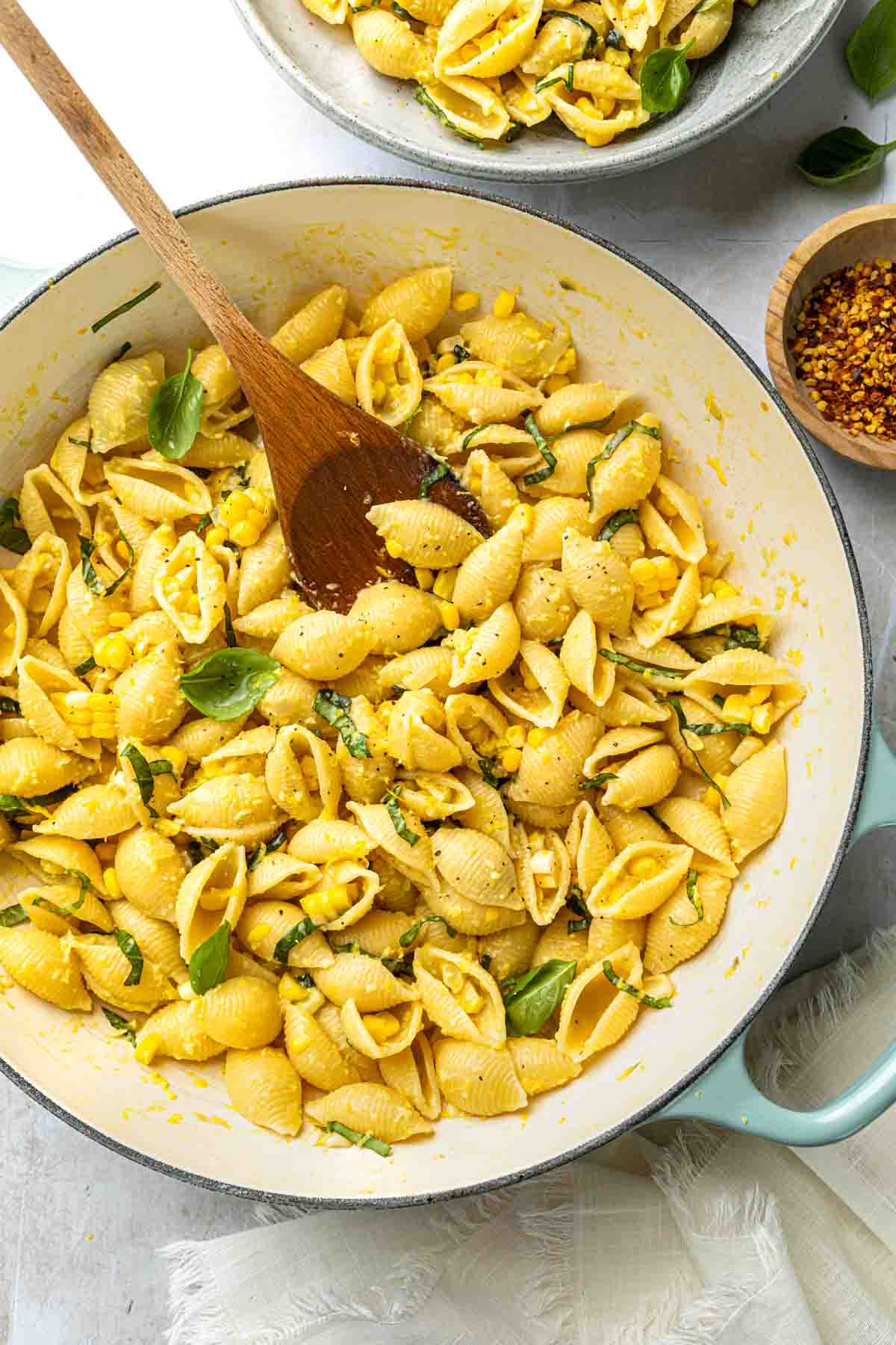 A large skillet with shell pasta, fresh corn sauce and sliced basil, with a wooden spoon. 