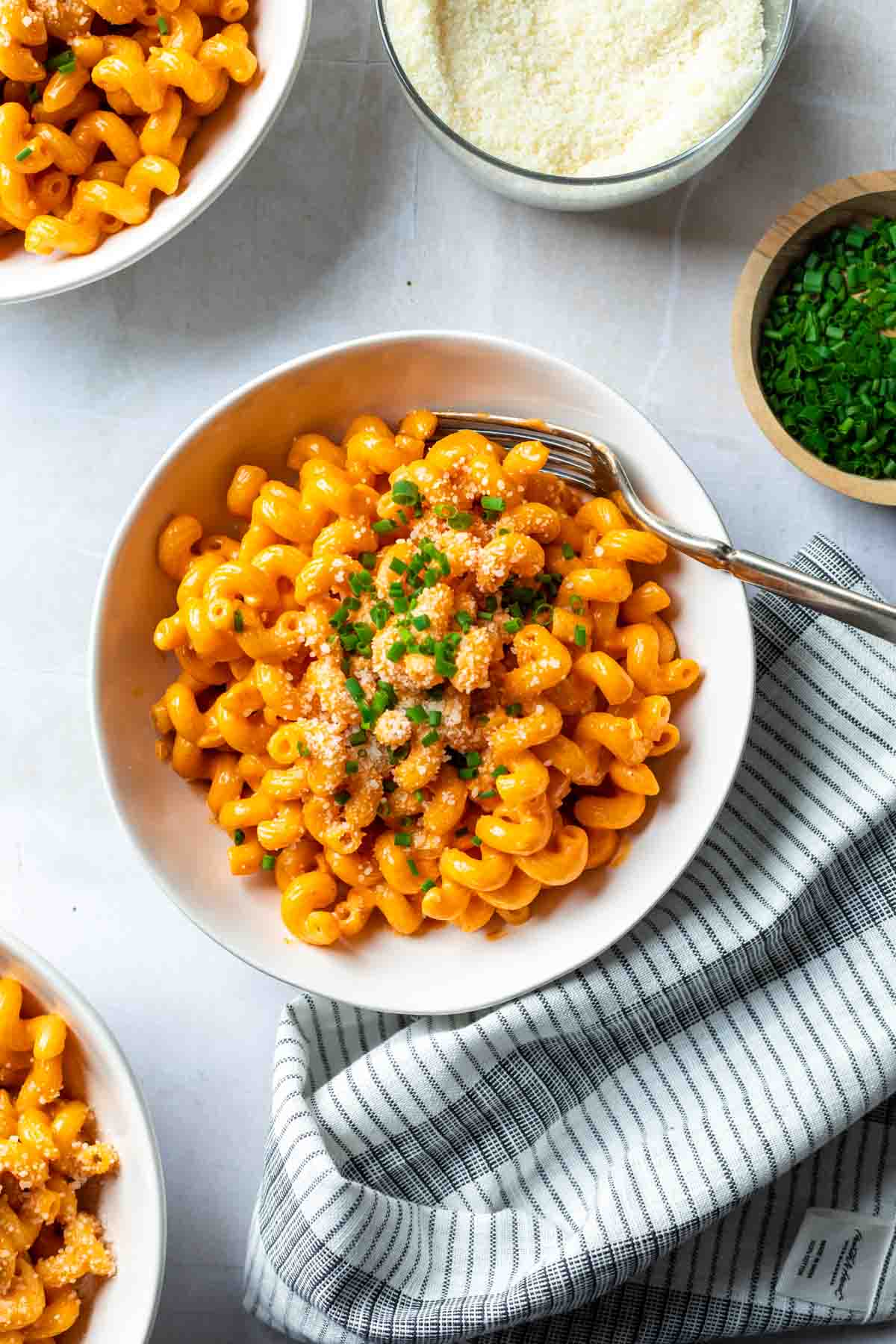 Bowls of cavatappi noodles garnished with grated cheese and chives, a bowl of chives to the side with a bowl of grated cheese. 