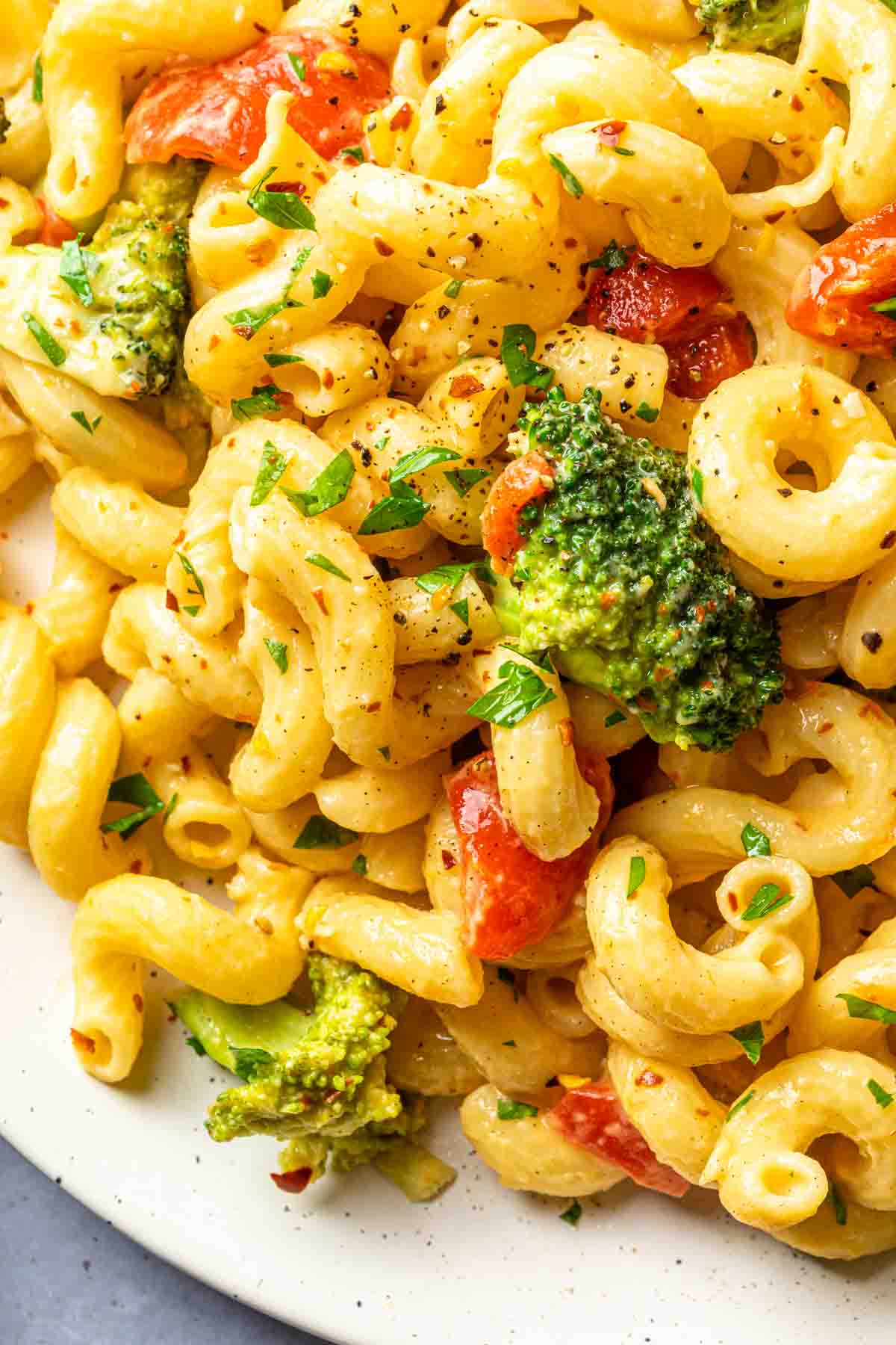 Close up of broccoli, roasted red peppers and cavatappi pasta on a plate, garnished with crushed red peppers and fresh parsley.