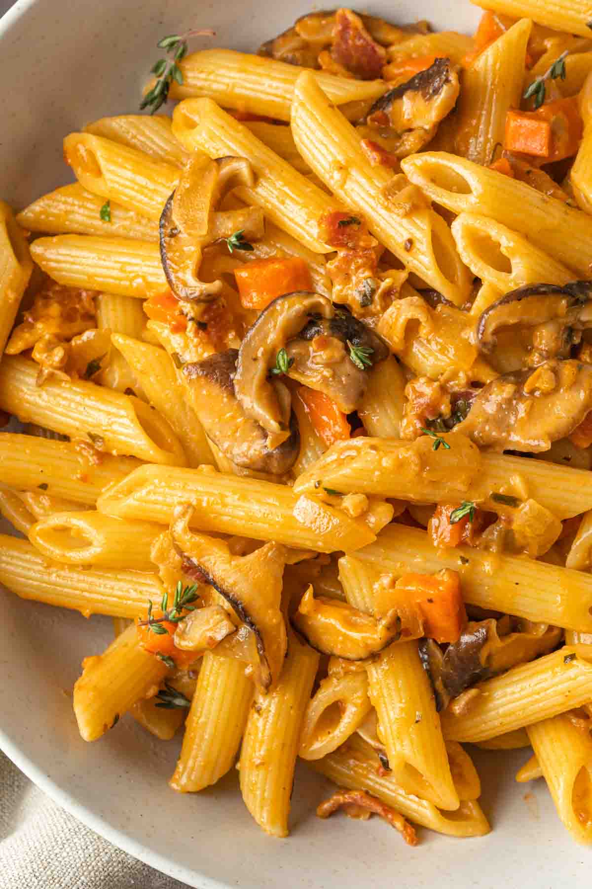 Close up of penne pasta with a mushroom sauce, showing the mushrooms, carrots, onions and fresh thyme in the sauce. 