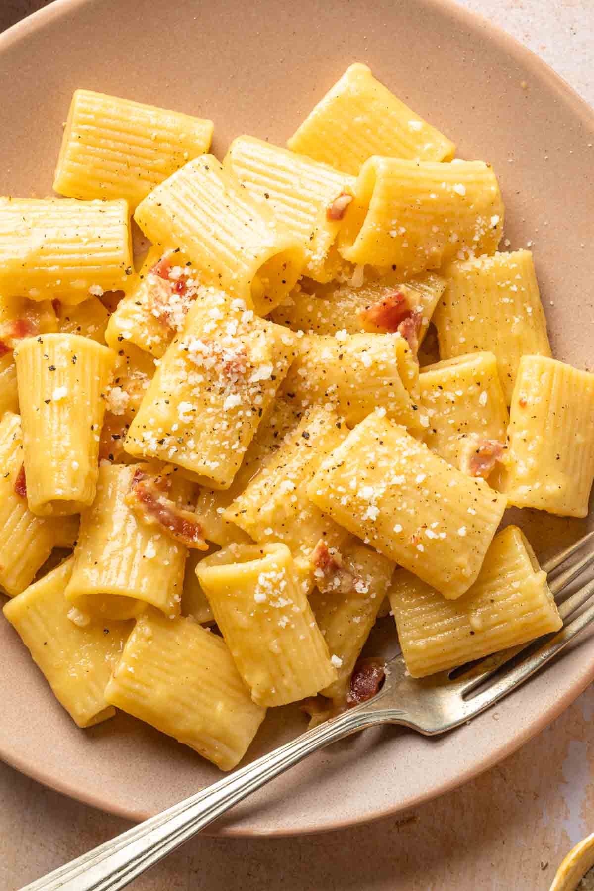 Close up of rigatoni carbonara in a bowl, garnished with grated cheese and cracked black pepper. 