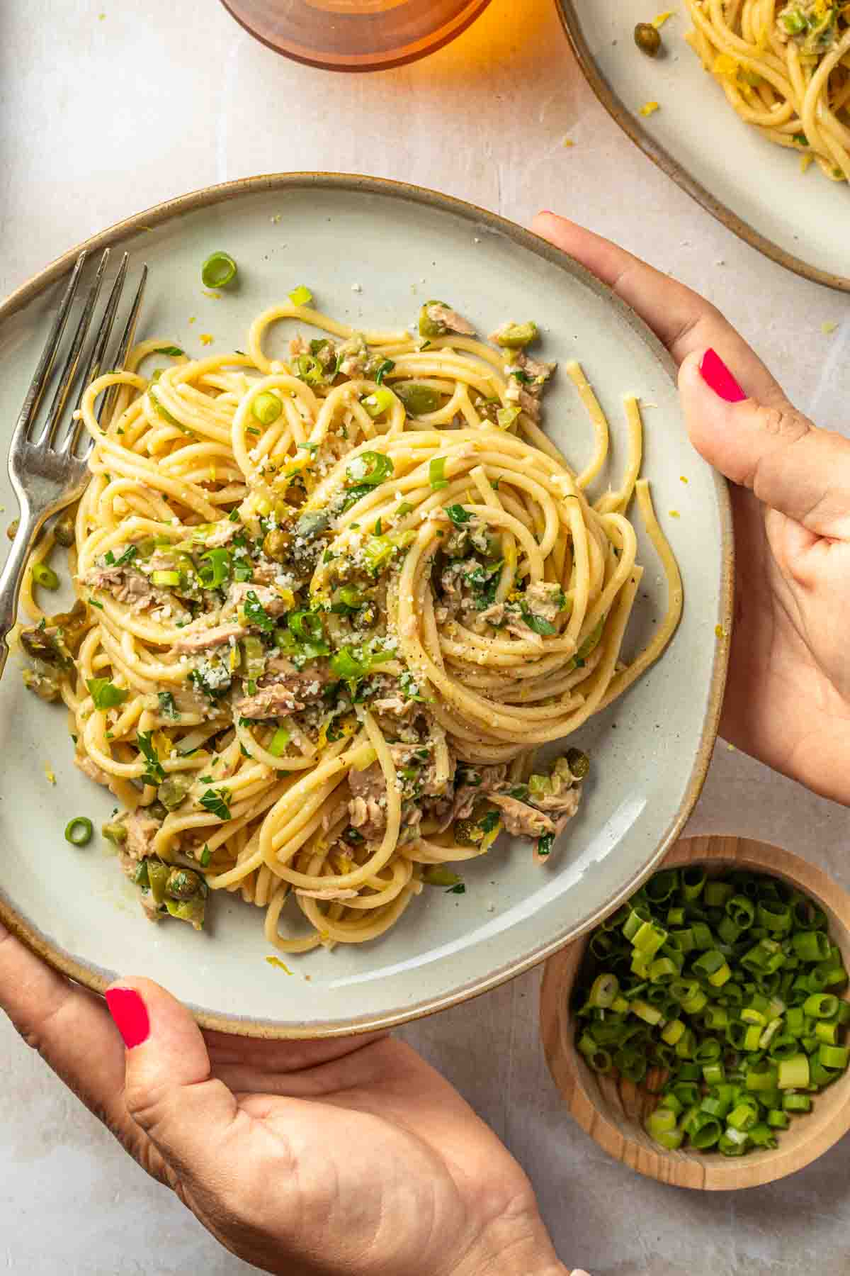 Hands holding a plate of pasta with capers, tuna, green onions and olives. 