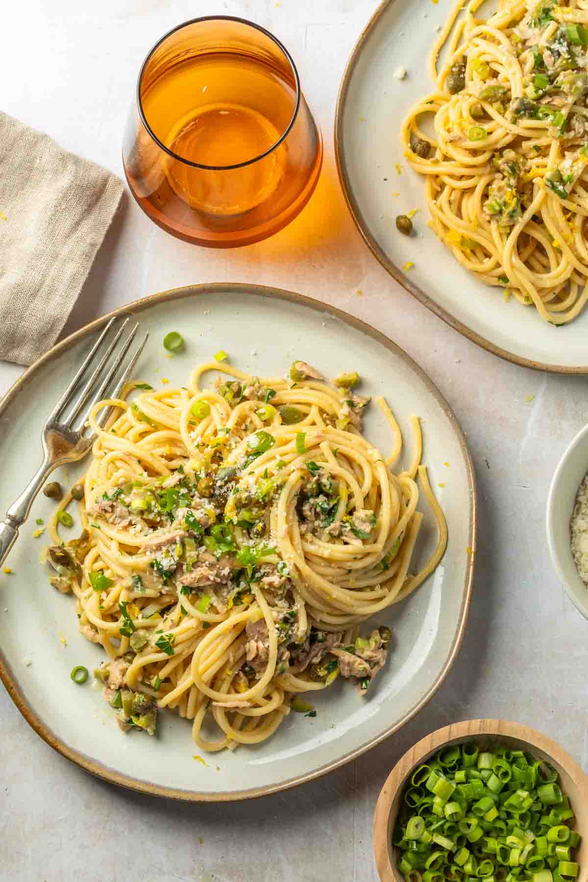 A couple of plates of pasta next to a bowl of grated pecorino romano cheese, a bowl of sliced green onions and a wine glass. 