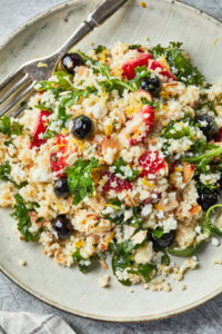 A plate of pastina salad with kale, blueberries, strawberries, crumbled feta cheese, toasted almonds, and lemon zest garnish, served with a fork.