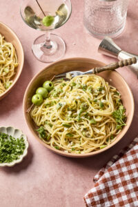 A bowl of spaghetti topped with chopped herbs, grated cheese, and three green olives sits on a pink table beside a glass of white wine, a small dish of herbs, and a red checkered cloth.