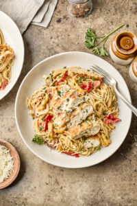 A plate of creamy pasta with grilled chicken strips, red and yellow bell peppers, and parsley, served on a rustic table with a fork, grated cheese, and condiments nearby.