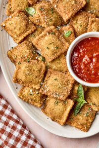 A plate of golden, breaded toasted ravioli garnished with herbs is served with a small bowl of marinara sauce. A red and white checkered napkin is beside the plate.