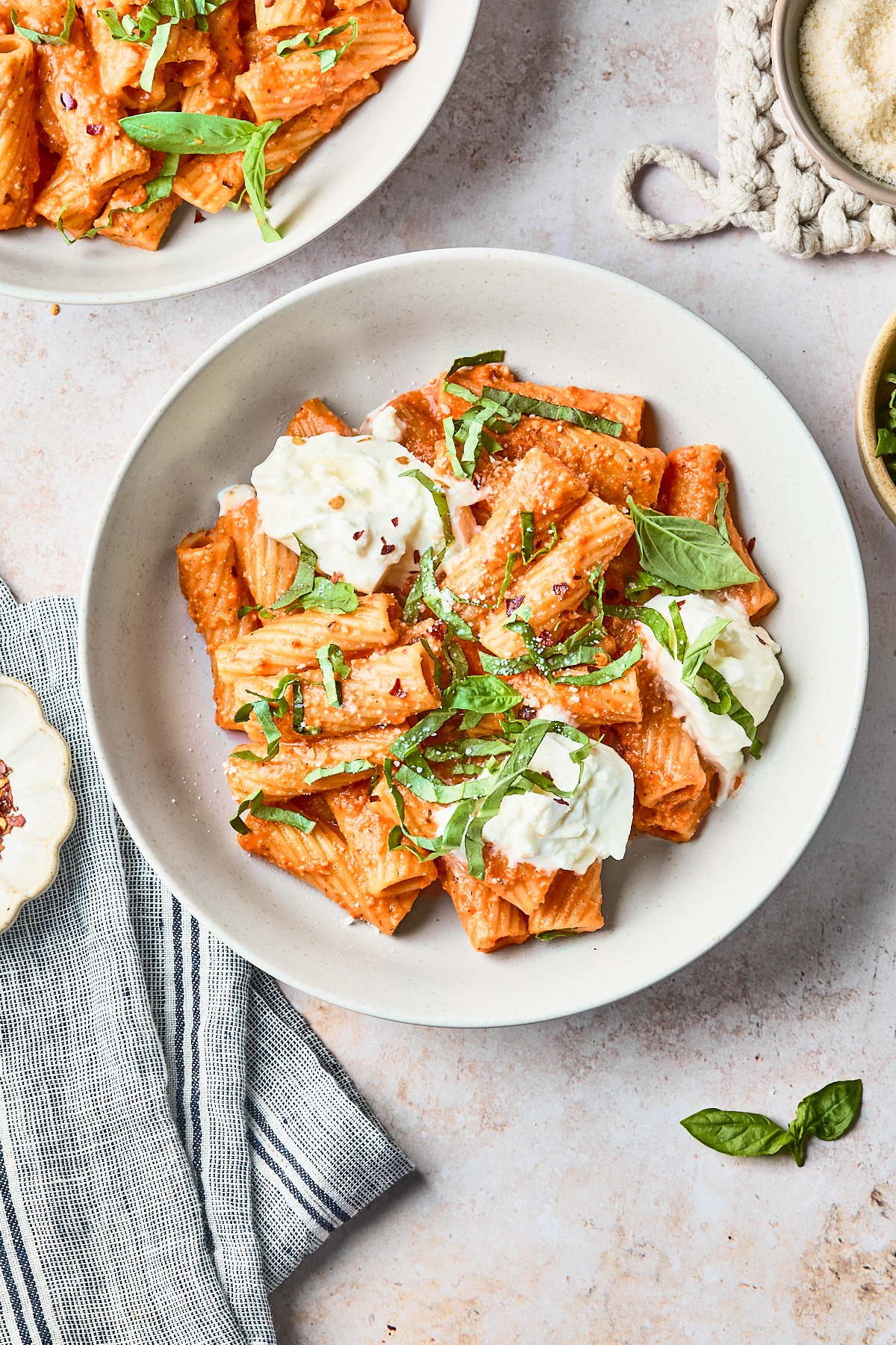 A white bowl of rigatoni pasta in creamy red sauce, topped with fresh basil and dollops of burrata cheese, sits on a light surface next to a striped cloth and a small bowl of grated cheese.