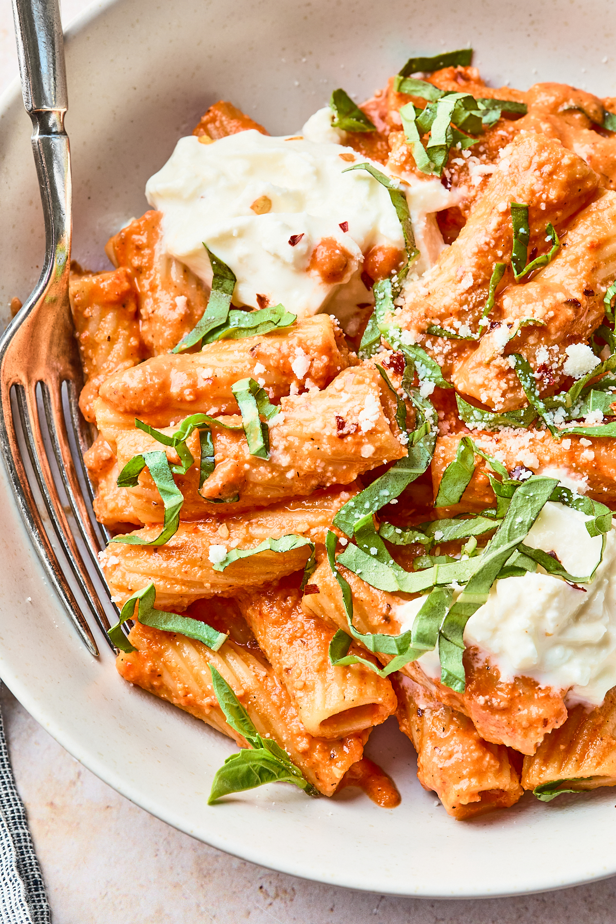 A plate of rigatoni pasta with creamy tomato sauce, topped with fresh basil, dollops of ricotta cheese, grated parmesan, and red pepper flakes, with a fork resting on the side.