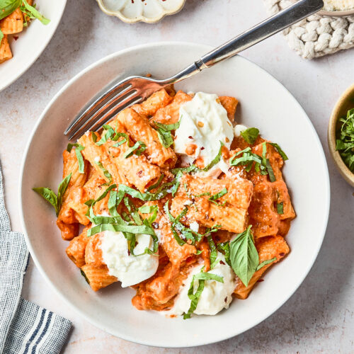 A bowl of rigatoni pasta in a creamy tomato sauce, topped with dollops of ricotta cheese, fresh basil, and grated parmesan, with a fork resting on the side. Surrounding dishes contain chili flakes and more cheese.