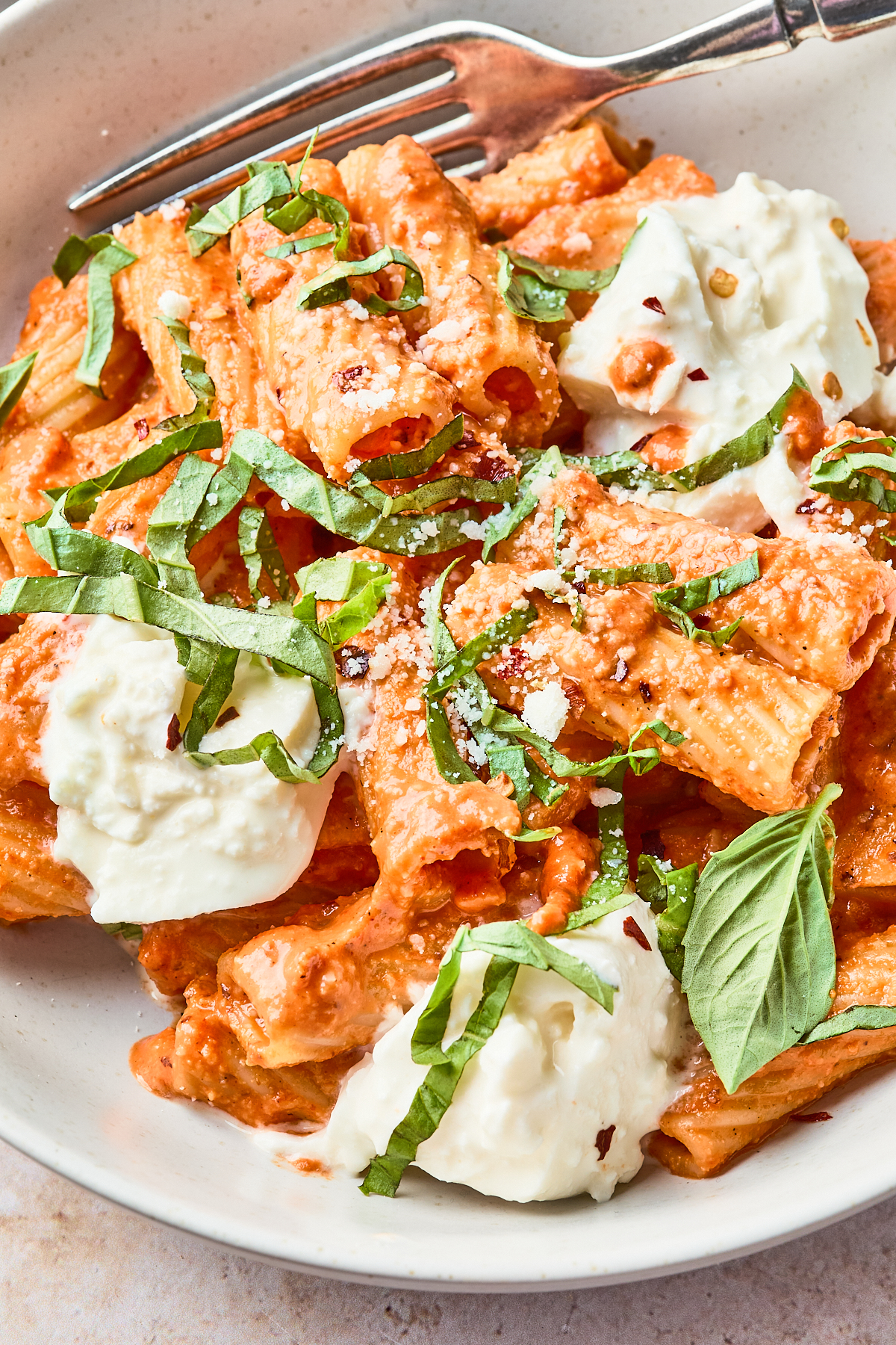 A bowl of rigatoni pasta in creamy tomato sauce, topped with dollops of ricotta cheese, grated parmesan, fresh basil ribbons, red pepper flakes, and garnished with basil leaves. A fork rests on the side of the bowl.