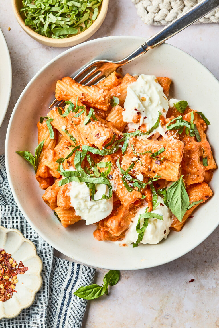 A bowl of rigatoni pasta in a creamy tomato sauce, topped with fresh mozzarella, grated cheese, and chopped basil, with a fork resting in the bowl. Small bowls of basil and red pepper flakes are nearby.