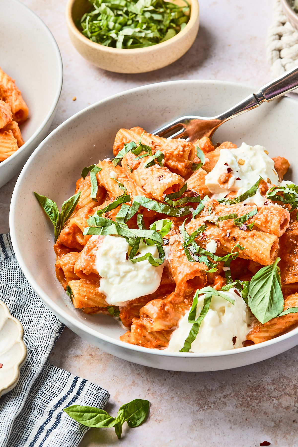 A bowl of rigatoni pasta in creamy tomato sauce, topped with dollops of ricotta cheese and fresh basil strips, with a fork resting in the bowl. A small bowl of chopped basil is in the background.